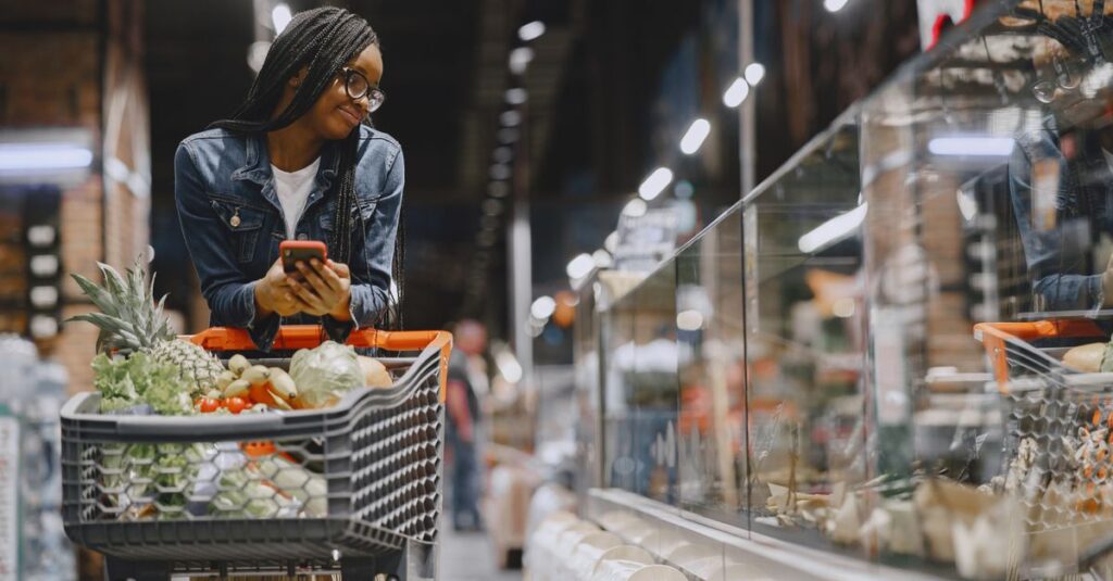 A young woman with afro braids grocery shopping in a supermarket, using a mobile phone.
